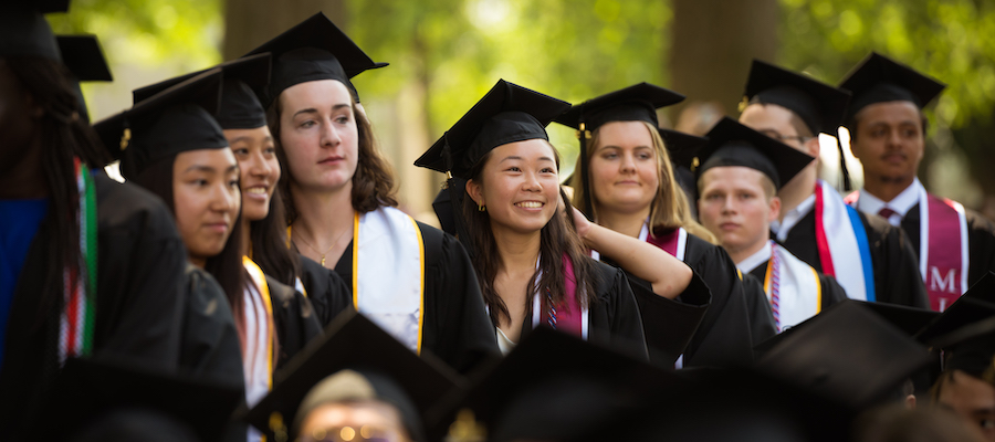 Image of graduates in line and smiling. They are all wearing a black gown and cap.