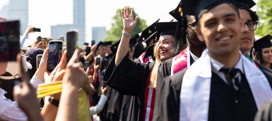 Image of the graduate division of the academic procession. One of the graduates in the image is waving towards the guests in the audience who are using their phones to take photos of the procession